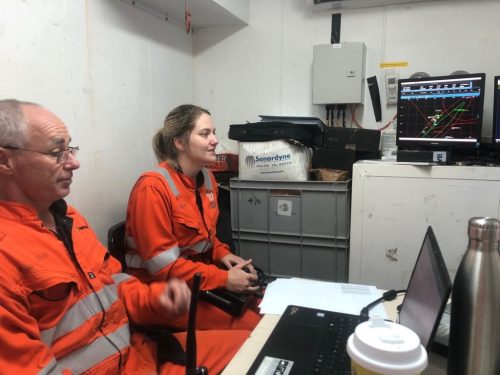 Two professionals in high-visibility orange overalls with reflective stripes are seated inside a control room with white walls. The individual on the left appears to be operating a joystick, while the person on the right, who seems to be in mid-conversation, looks on. They are surrounded by work-related paraphernalia, including a monitor displaying a graph with multiple coloured lines, suggesting some form of data monitoring or analysis. Personal items such as a stainless steel bottle and a takeaway coffee cup, along with a laptop and operational equipment, are also visible on the desk, adding to the candid and occupied atmosphere of the scene.