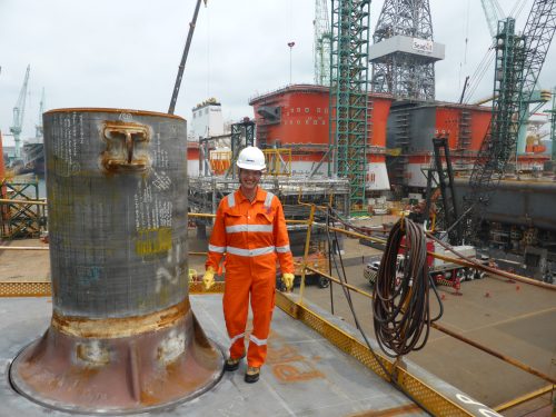 An individual in orange safety coveralls and a white helmet stands smiling on a shipyard platform beside a large metallic structure with visible writing and diagrams. In the background is a view of industrial, maritime construction with multiple cranes, a docked ship, and a large offshore platform under maintenance. Safety barriers and construction materials are scattered throughout the site.