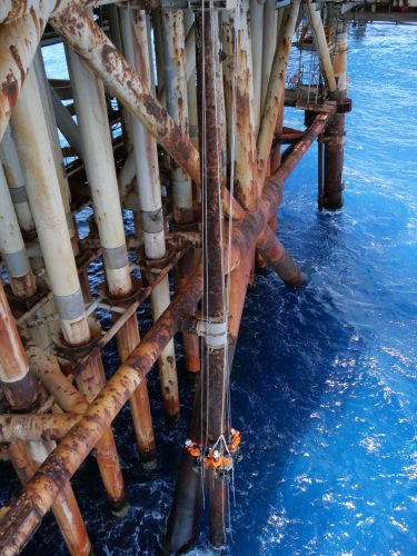 Workers in orange safety gear perform maintenance on an offshore rig's structure. They are attached to a vertical buoyancy aid amidst rusty pipes and beams. The sea below is a deep blue.