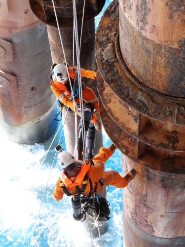 Two workers in orange safety suits and white helmets are suspended on ropes beside an offshore platform's massive, rusted cylindrical legs. They are equipped with safety harnesses and gear, with the ocean visible below.