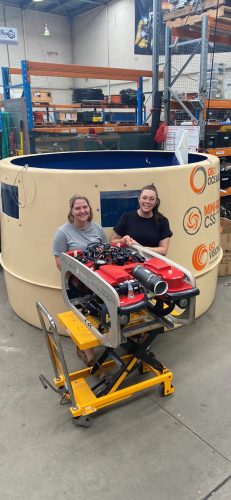 Two individuals are smiling at the camera, seated behind a red and black Remotely Operated Vehicle (ROV) on a yellow lift. They are in a warehouse with storage racks and equipment in the background. The large cylindrical object behind them features logos, including "GEO OCEANS" and "VERTTECH GROUP".