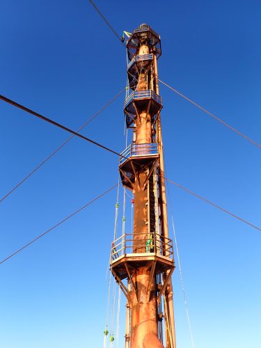 A towering metal structure with platforms, ladders, and safety railings rises against a clear blue sky. The structure, exhibiting a weathered rust patina, is equipped with pulleys and cables, indicative of its use for lifting or adjusting heavy equipment. The perspective is from the ground, looking up the structure's full height, emphasising its significant elevation and the work's verticality.